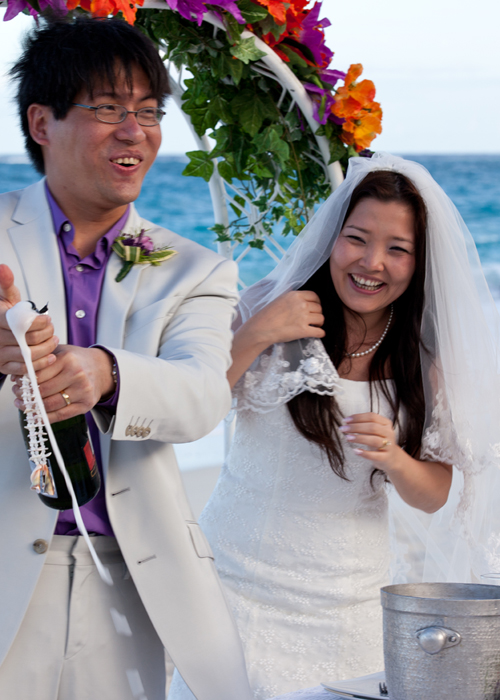 Newlyweds laughing as the groom opens champagne after their beach ceremony